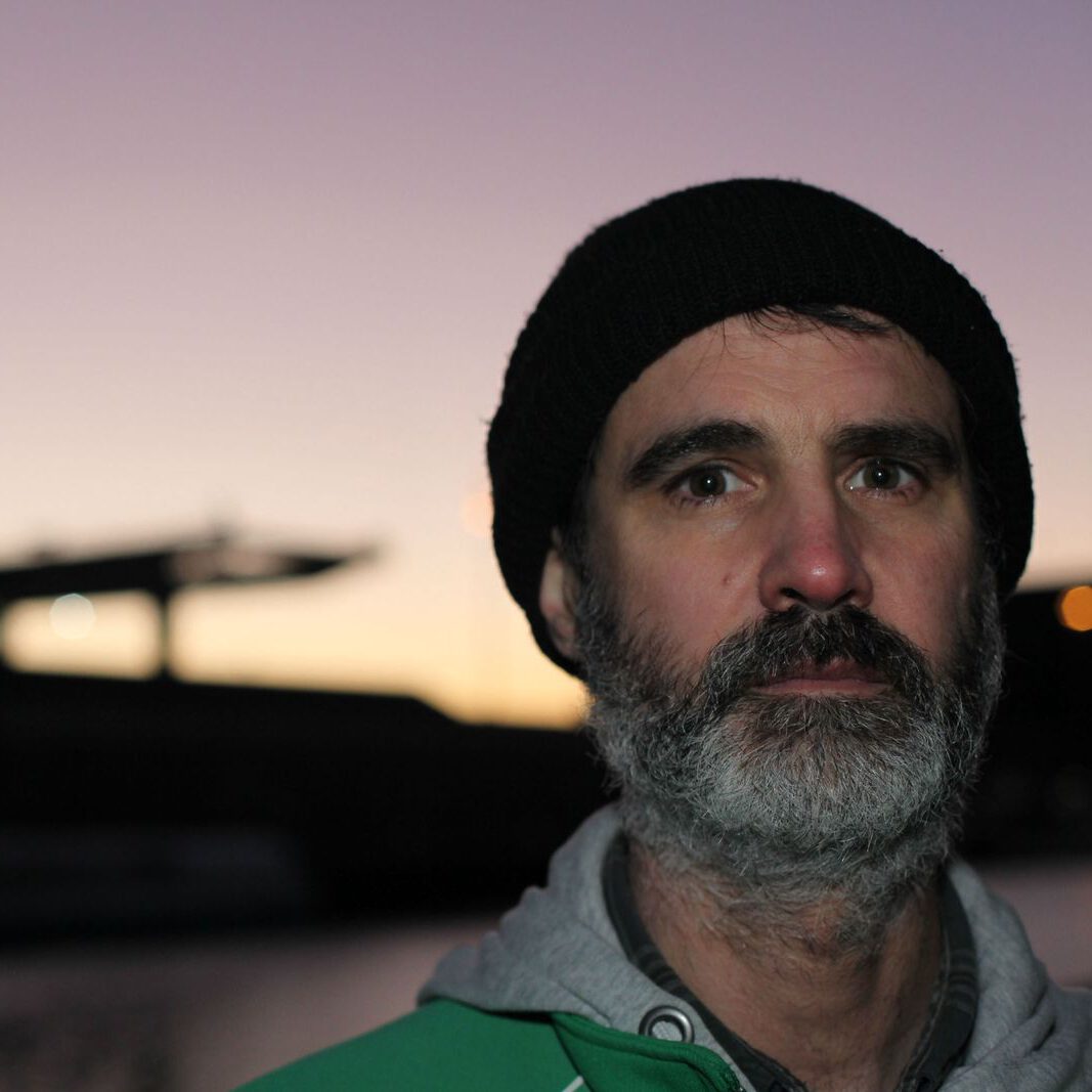 Portraitfoto vom Künstler "Oldseed". Er trägt Bart, eune schwarze Wollmütze, einen grauen Hoodie und eine grüne Trainingsjacke. Im Hintergrund erkennt man schemenhaft ein Stadion im Sonnenuntergang.