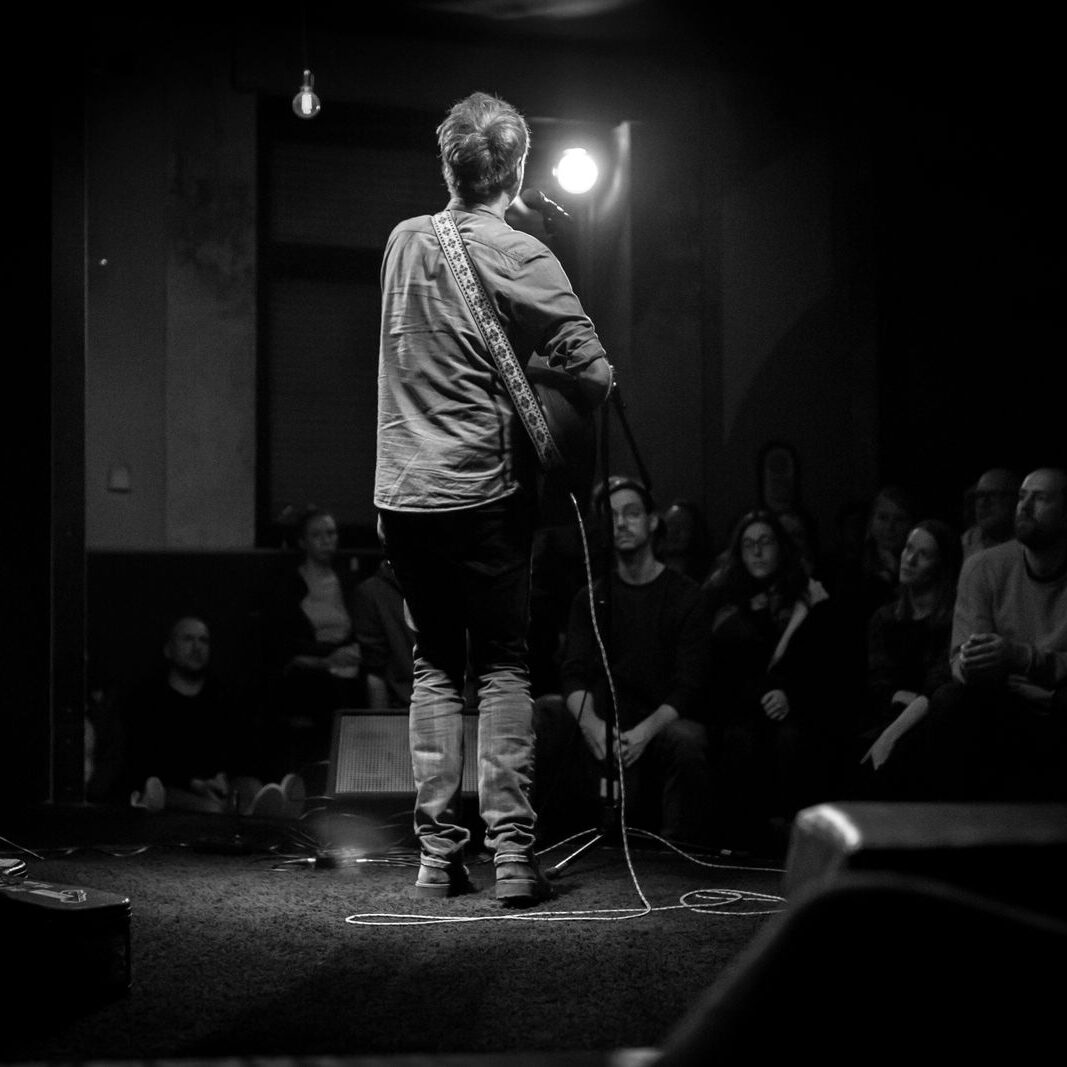 Schwarz-Weiß-Fotografie vom Musiker "Oldseed". Man sieht ihn von Hinten mit einer Gitarre vor sitzendem Publikum.